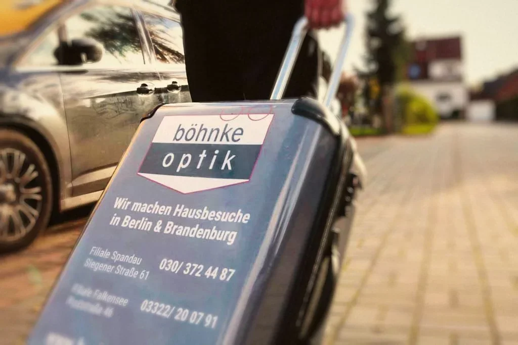 Ein Koffer mit dem Böhnke-Optik-Logo und der Aufschrift „Wir machen Hausbesuche in Berlin & Brandenburg“ wird über einen Gehweg gezogen.