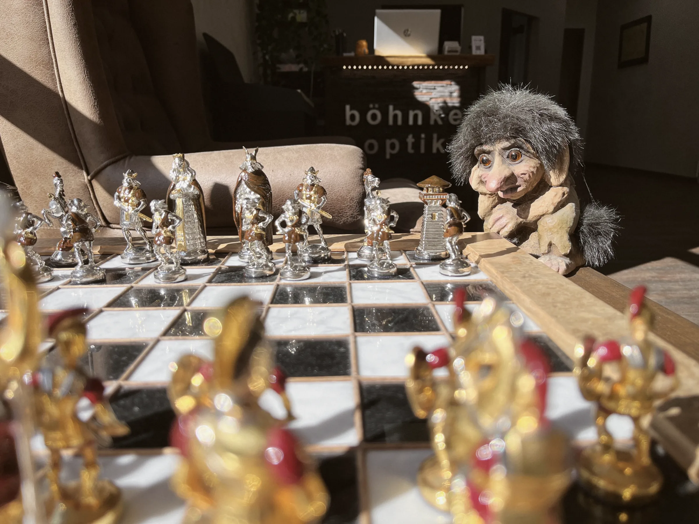 Ein Schachspiel mit goldenen und silbernen Figuren im Laden, dahinter ein Troll mit Brille und der Schriftzug Böhnke Optik.